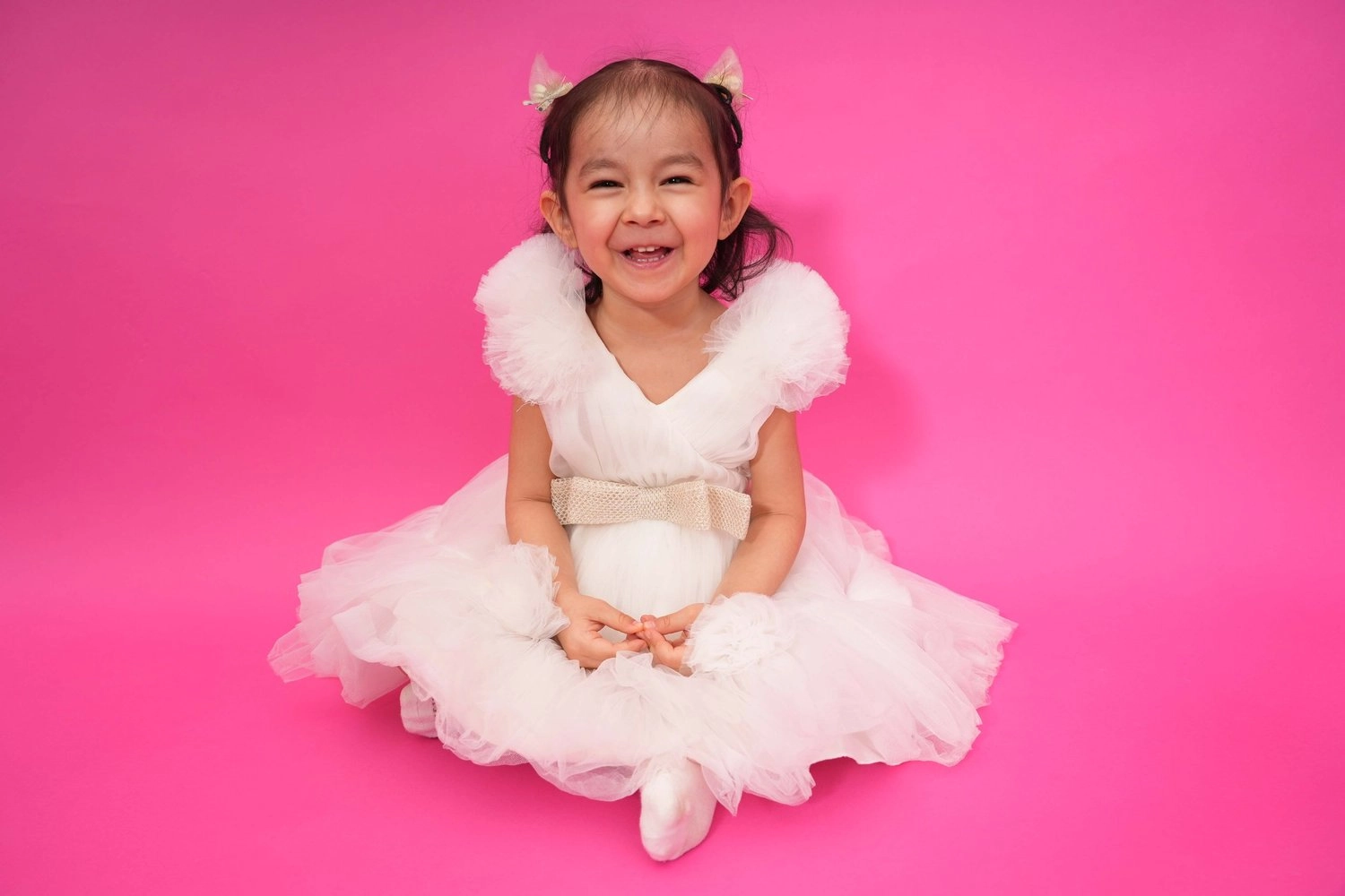 Ein lachendes Kind im weißen Kleid sitzt vor kräftigem Pink.