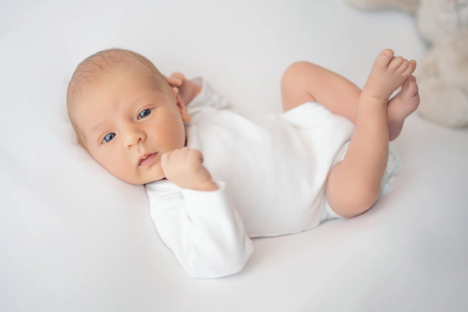 Ein waches Baby im weißen Body liegt mit angehobenen Füßen.