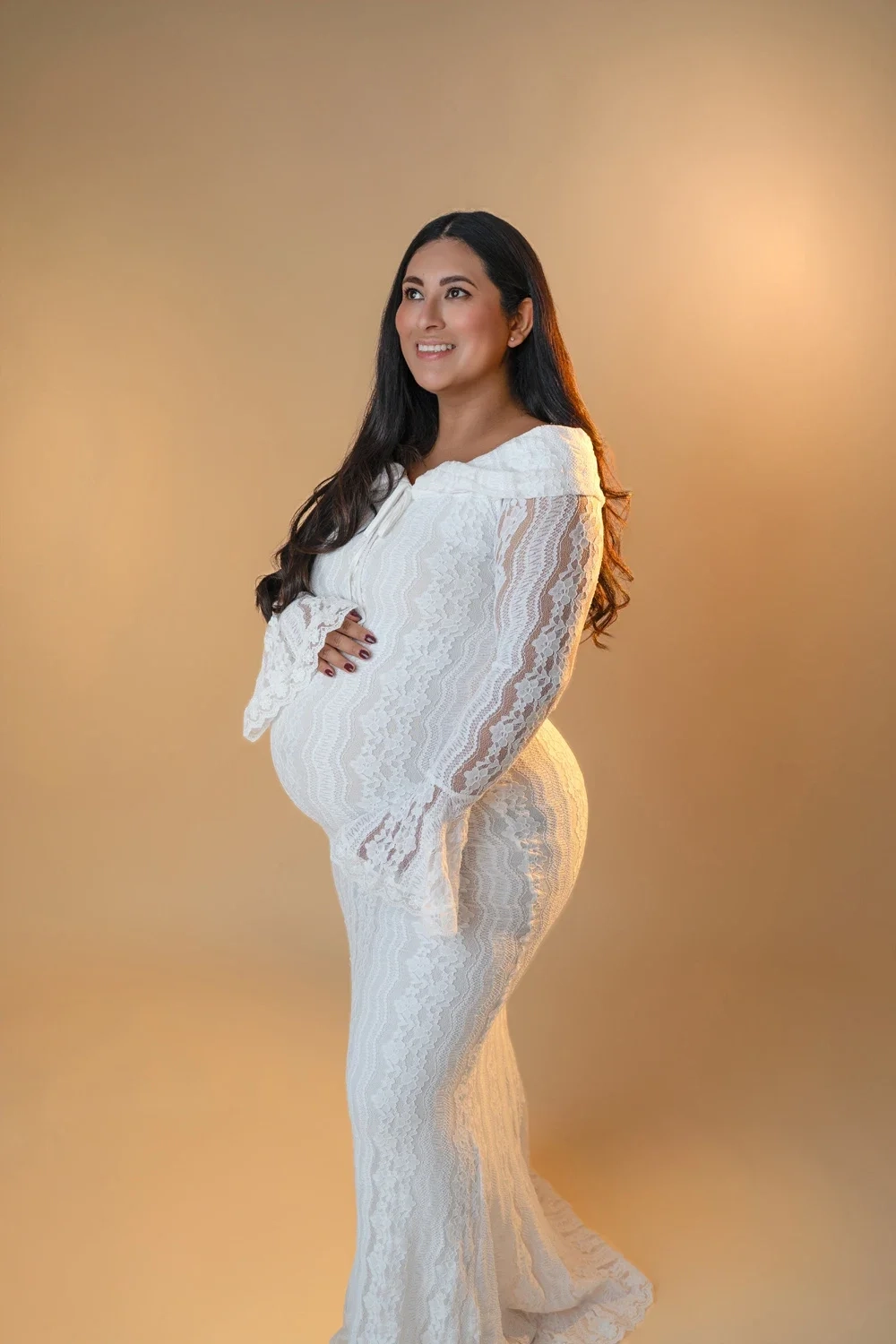 Eine schwangere Frau im weißen Spitzenkleid hält lächelnd ihren Bauch.