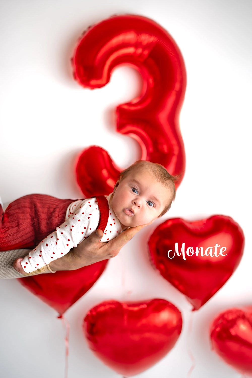 Ein Baby liegt zwischen roten Herzballons neben einer großen Drei.