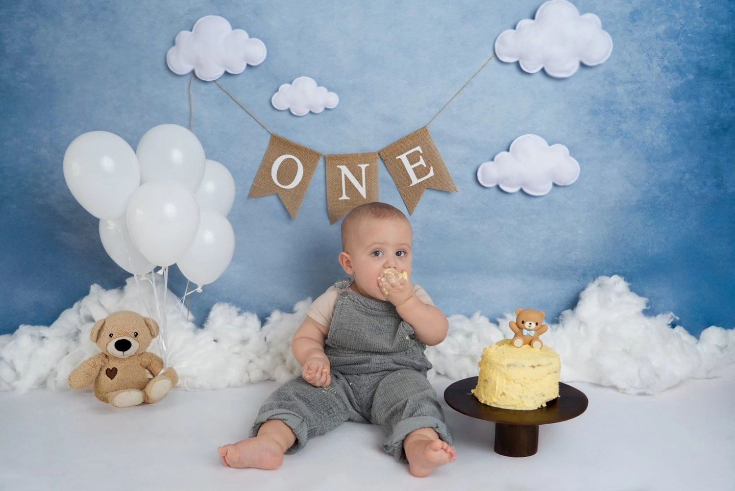 Ein sitzendes Baby probiert Torte vor blauer Wolkendekoration und Luftballons.