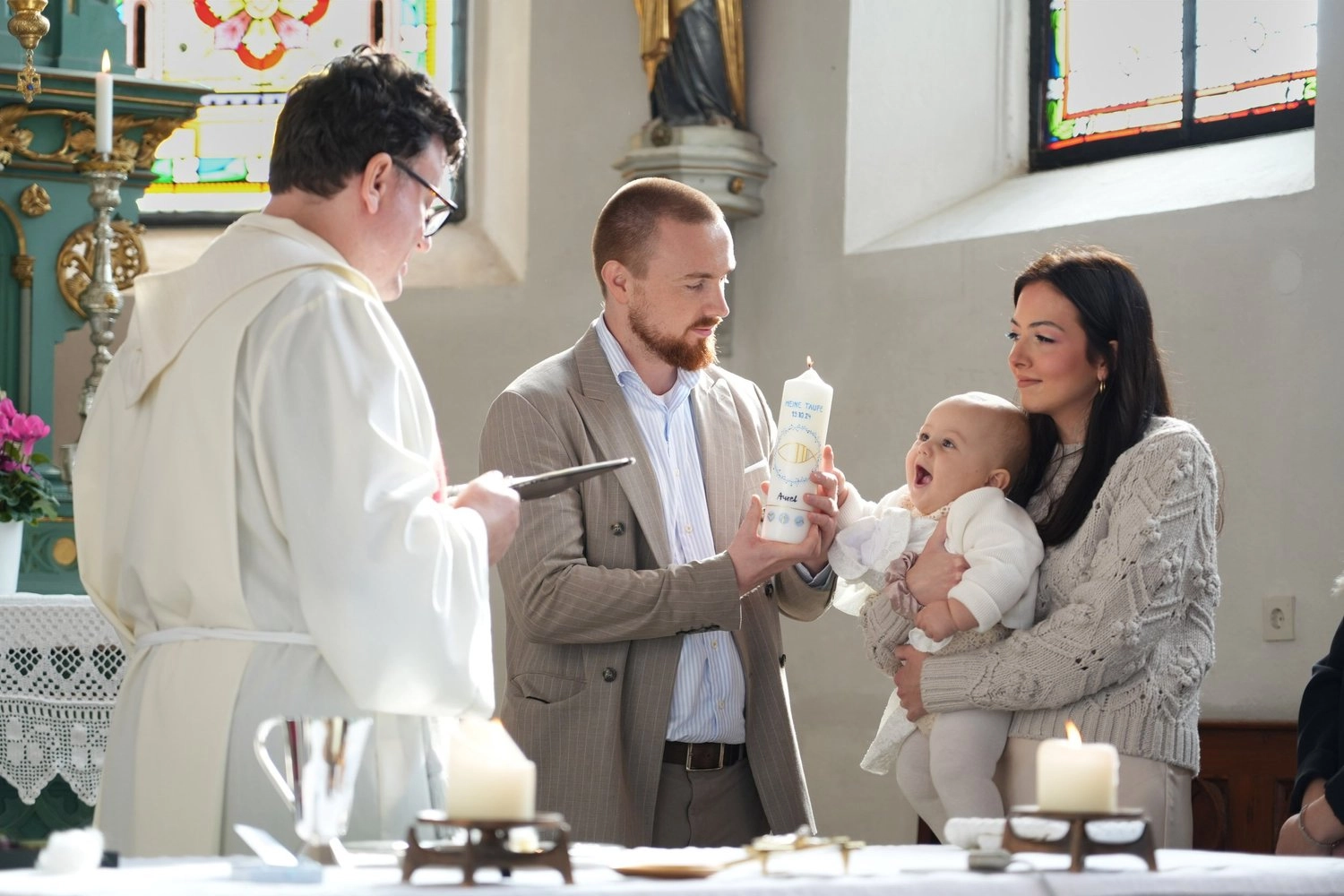 Ein Pfarrer begleitet eine Familie mit Baby am geschmückten Taufbecken.
