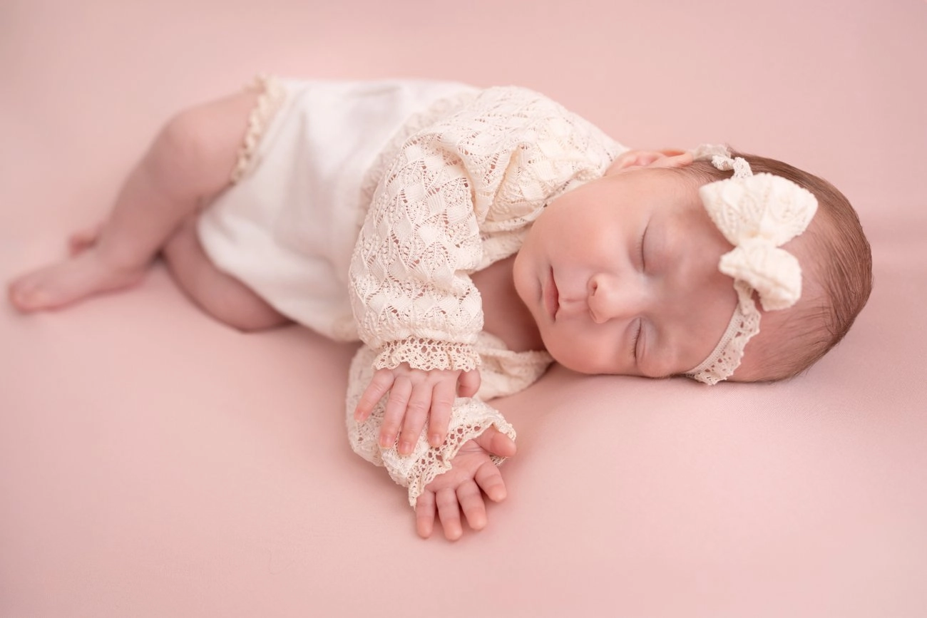 Ein schlafendes Baby mit Schleifenhaarband liegt auf einem rosa Untergrund.