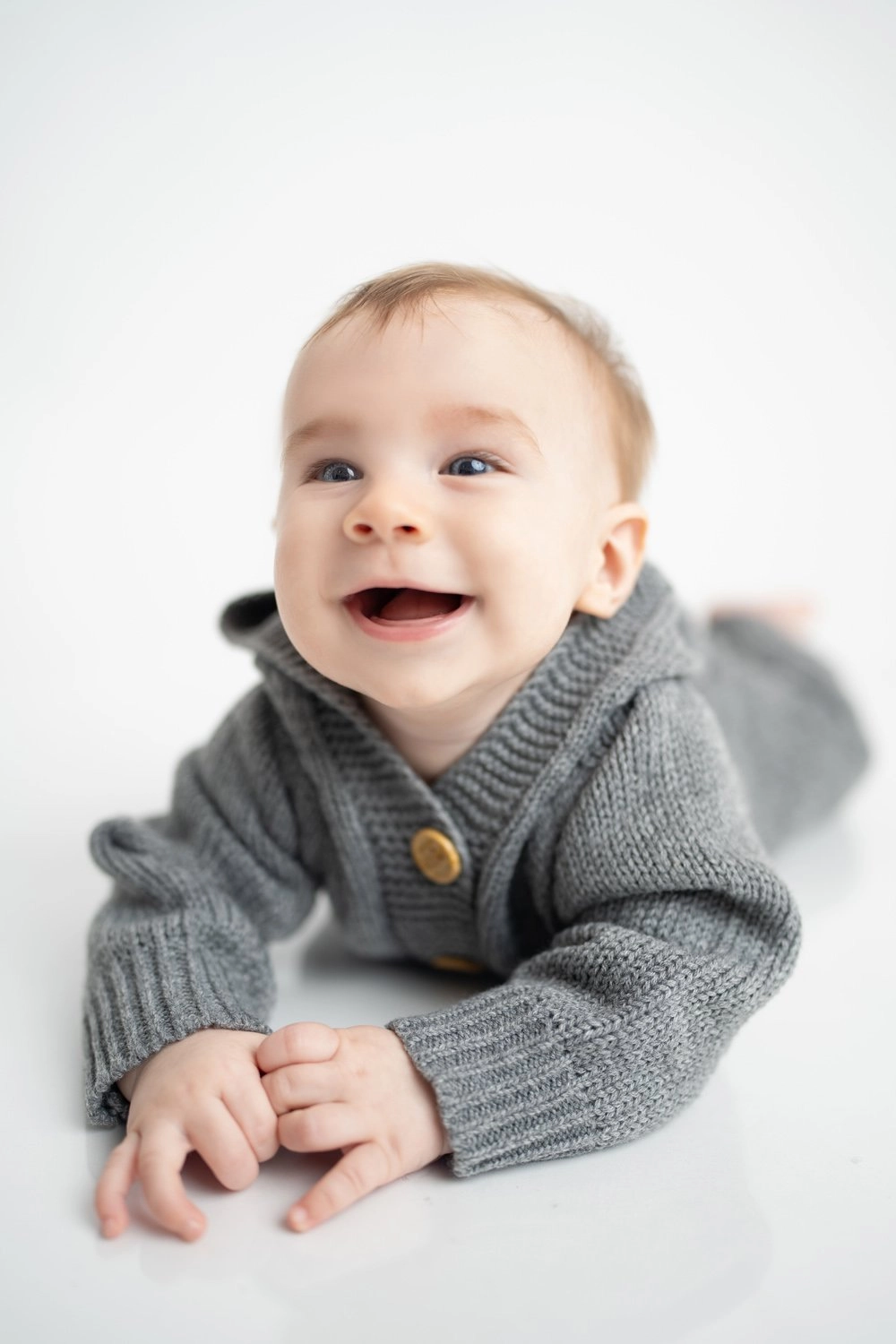 Ein lachendes Baby liegt im grauen Strickpullover auf hellem Untergrund.