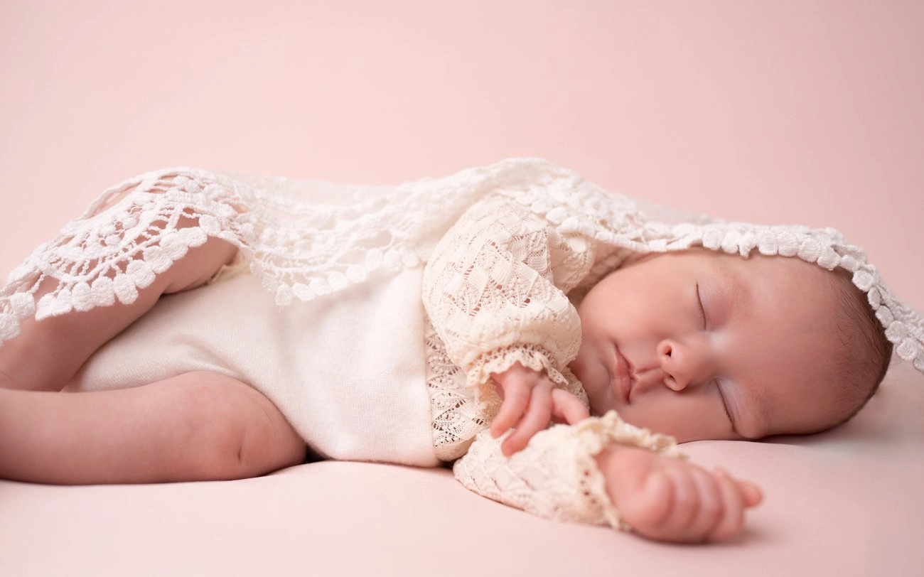 Ein schlafendes Baby in heller Spitze ruht seitlich auf rosa Untergrund.