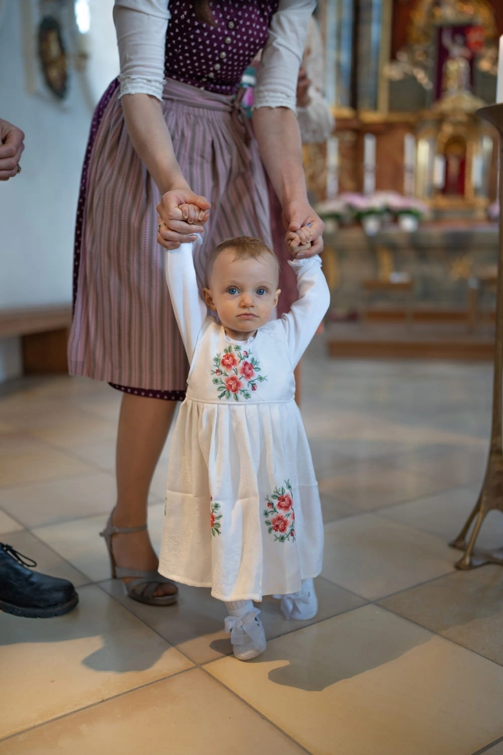 Ein kleines Kind im weißen Kleid steht begleitet in einer Kirche.