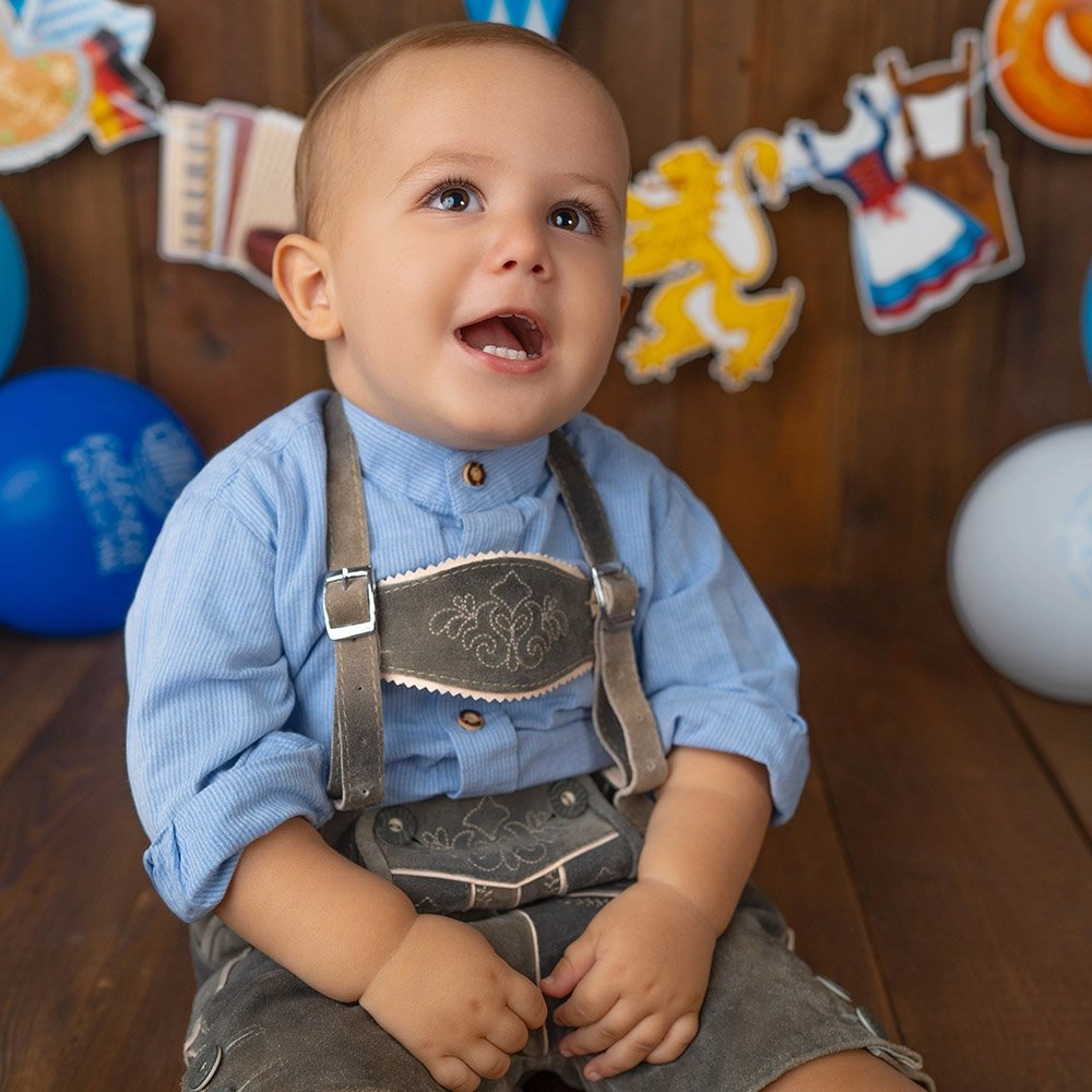 Ein lachendes Baby trägt blaue Tracht vor einer bunten Festgirlande.