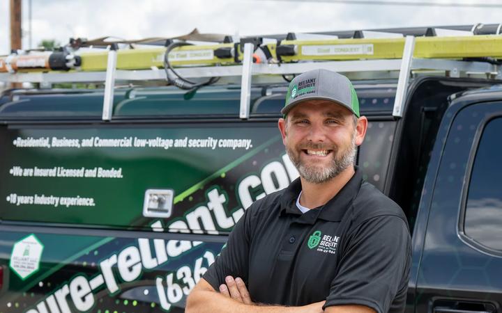 A Reliant Security technician beside a branded service truck loaded with ladders.