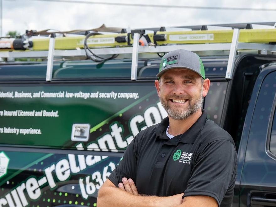 A Reliant Security technician beside a branded service truck loaded with ladders.