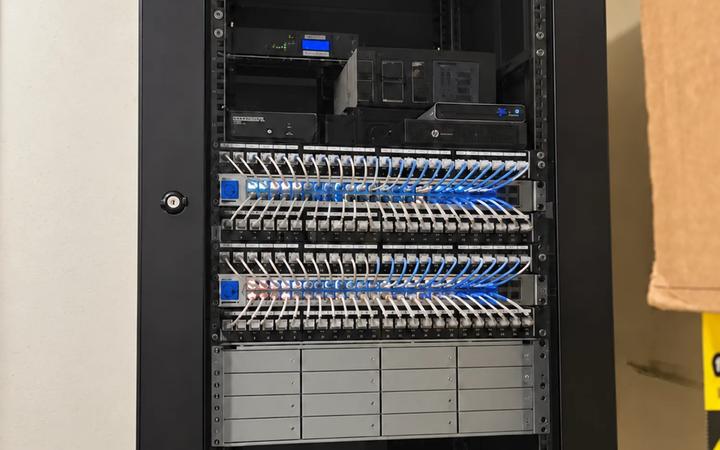 A wall-mounted network enclosure with patch panels, blue network runs, and a UPS.