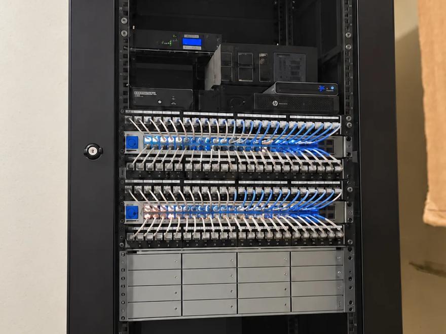 A wall-mounted network enclosure with patch panels, blue network runs, and a UPS.