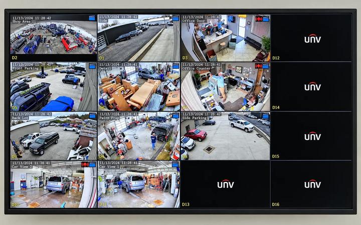 A wall-mounted monitor showing a sixteen-camera surveillance grid covering a shop floor, offices, and parking areas.