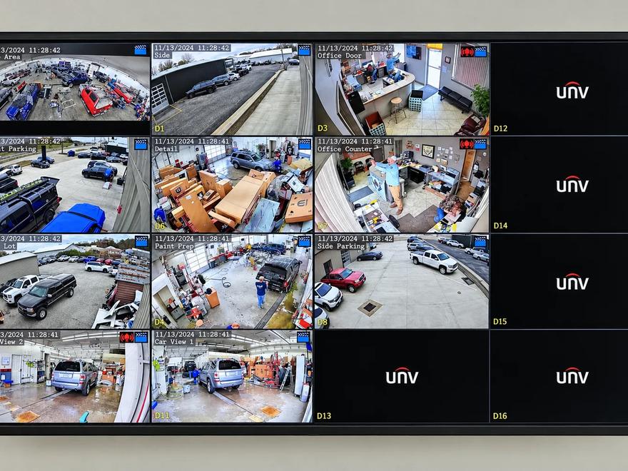 A wall-mounted monitor showing a sixteen-camera surveillance grid covering a shop floor, offices, and parking areas.