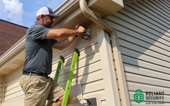 A Reliant Security technician on a ladder mounting a device under the eaves of a house.
