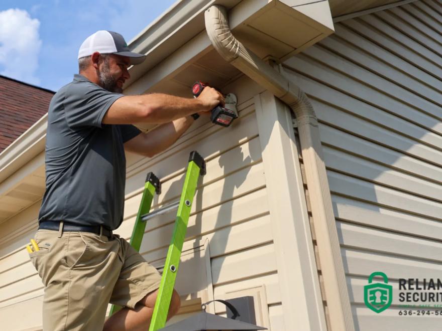 A Reliant Security technician on a ladder mounting a device under the eaves of a house.