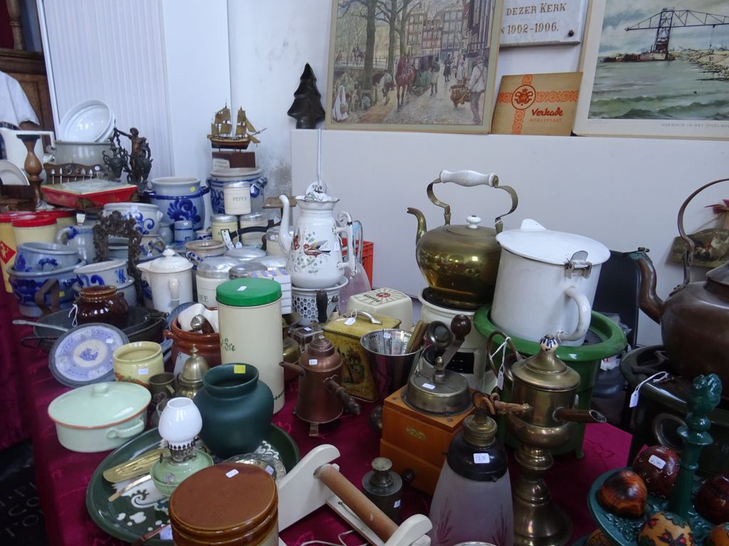 Tafel met koperen ketels, emaille pannen, steengoed en een oude lamp voor een witte muur