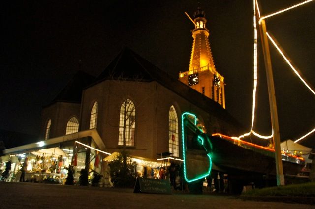 Verlichte kerktoren en marktkramen in de avond, naast een schip met lichtsnoeren