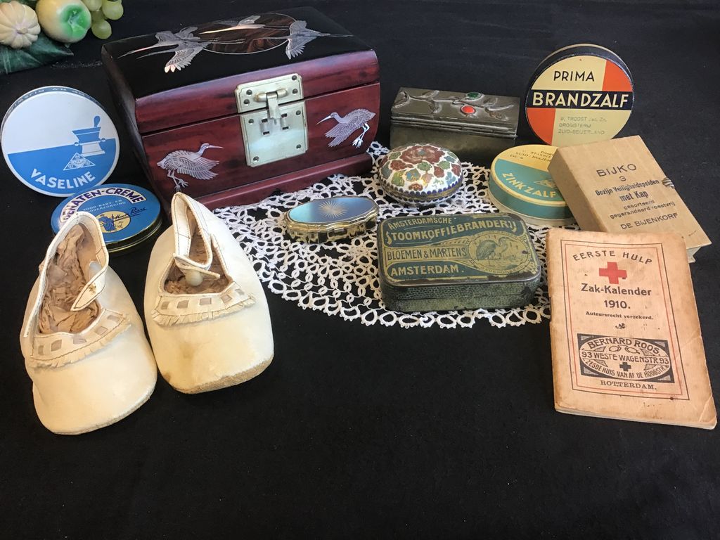 Baby shoes, a Chinese box, old tins and a pocket calendar from 1910 on a black cloth with lace