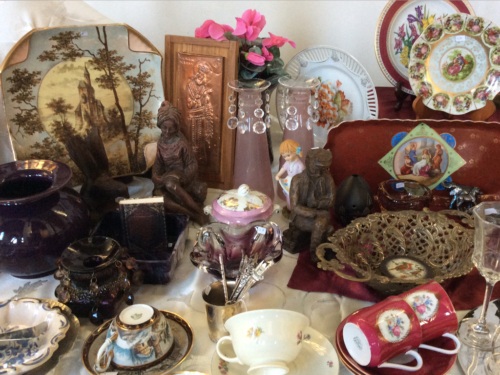 Table full of antiques: plate with a small church, copper relief, pink vases, bonbon dish and cups on a lace cloth