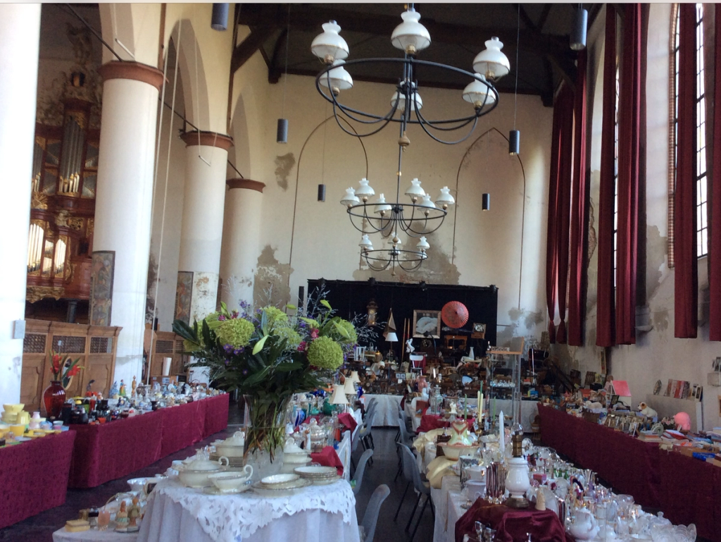 Church hall with long tables full of crockery and trinkets, burgundy cloths, a flower arrangement and chandeliers