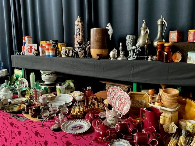 Tafel met rode kannen en kopjes, borden met roze motief en houten schalen, met tinnen kannen en bierpul op een plank erboven