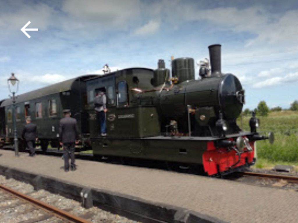 Zwarte stoomlocomotief met groen rijtuig en rode buffer op een perron, met twee spoorwegmedewerkers