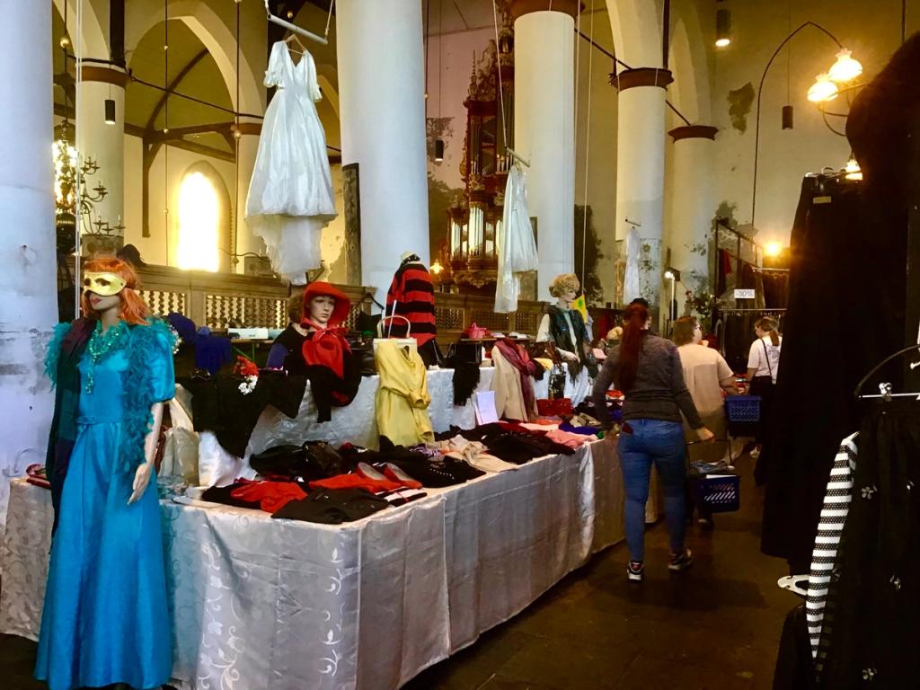 Kraam met kleding en een trouwjurk aan een pilaar, met bezoekers in de kerk