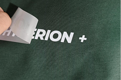 White heat-transfer logo film being applied to dark green fabric for custom drawstring bags