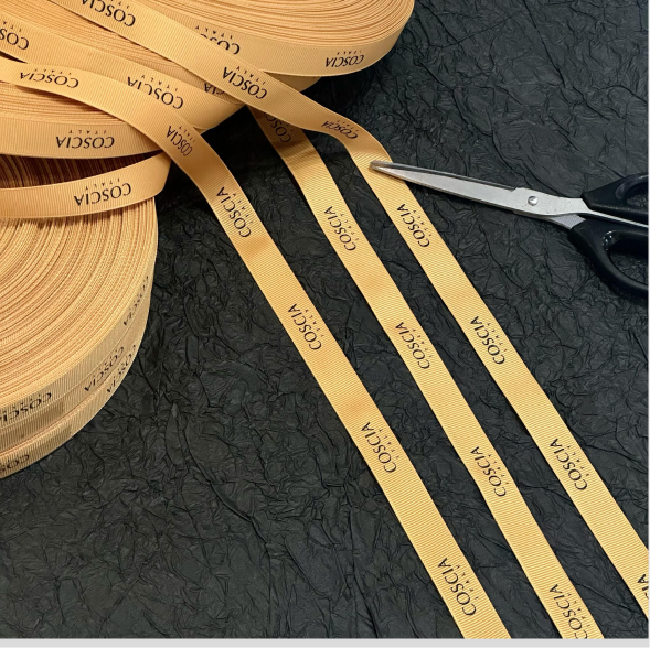 Three narrow warm amber ribbon strips with repeated COSCIA logos beside scissors