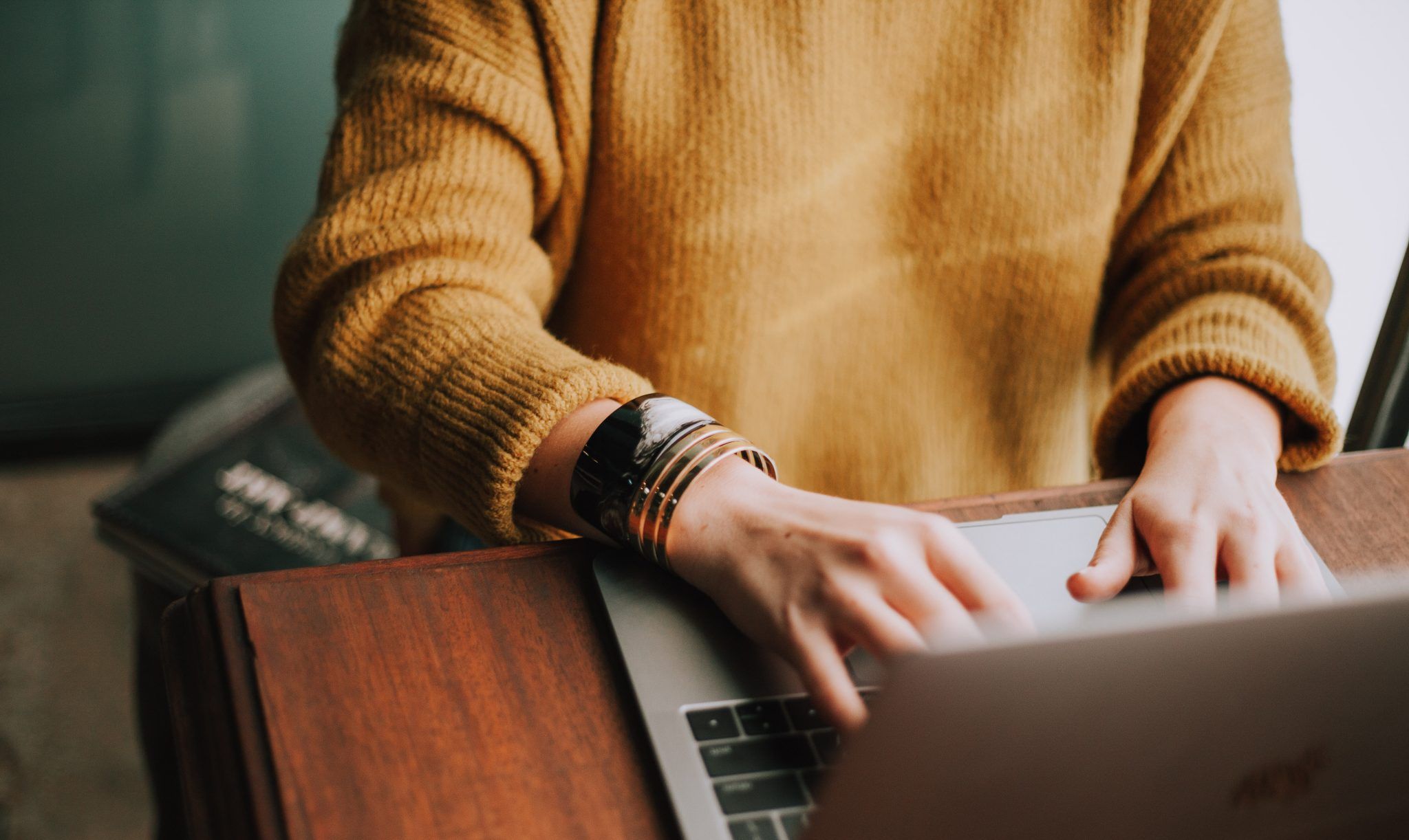 Person wearing a yellow sweater working on a laptop at a wooden desk