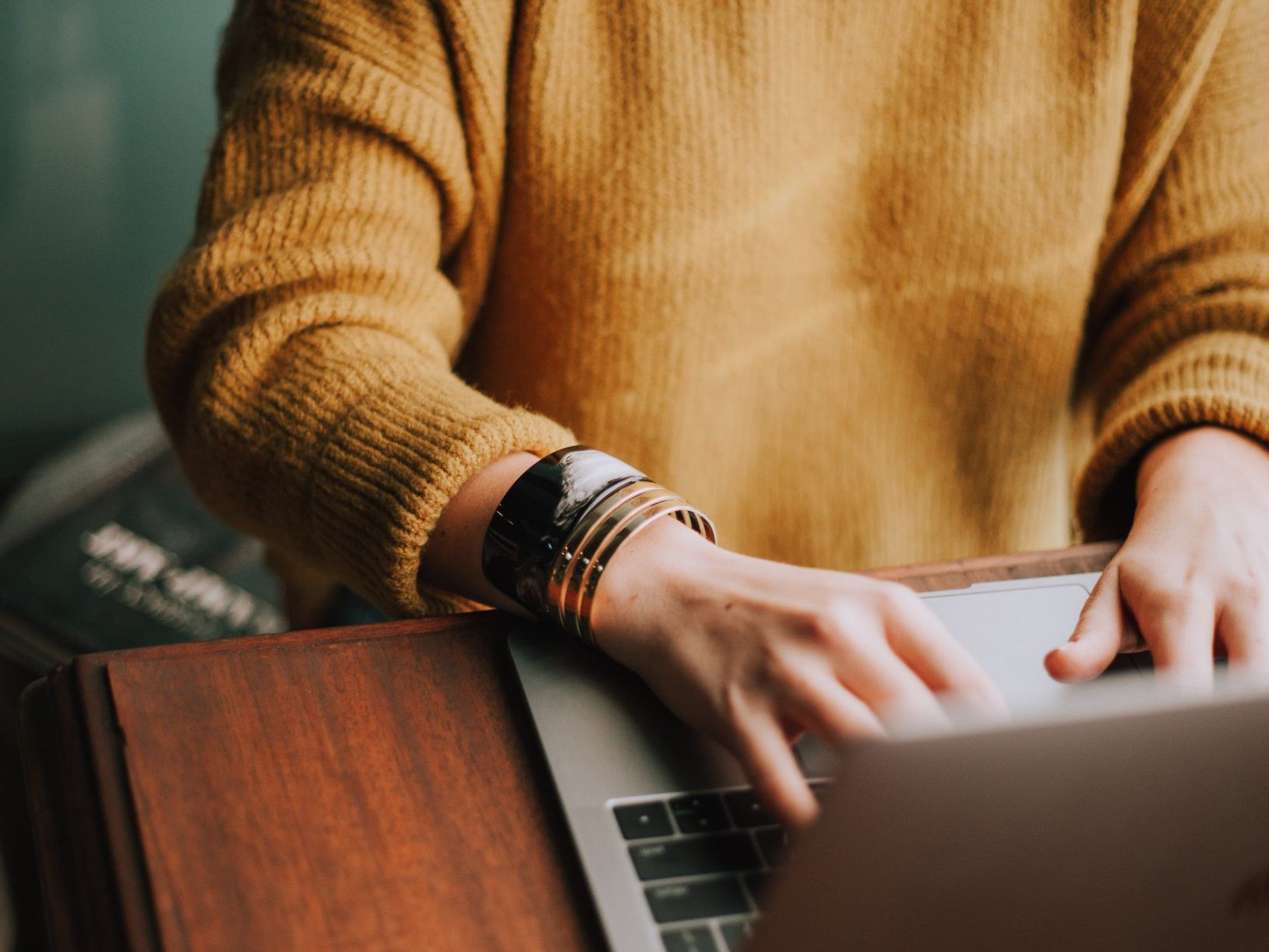 Person wearing a yellow sweater working on a laptop at a wooden desk