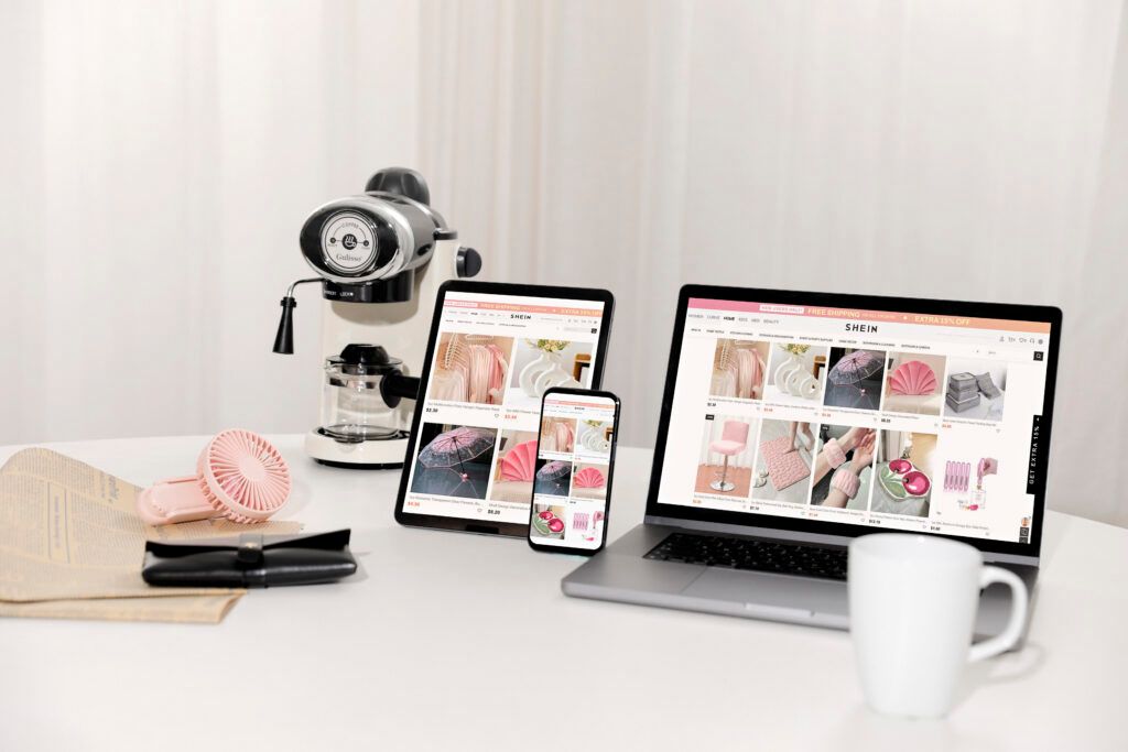 A desk setup with a coffee machine, a fan, a mug, and multiple devices (laptop, tablet, and phone) displaying the SHEIN website