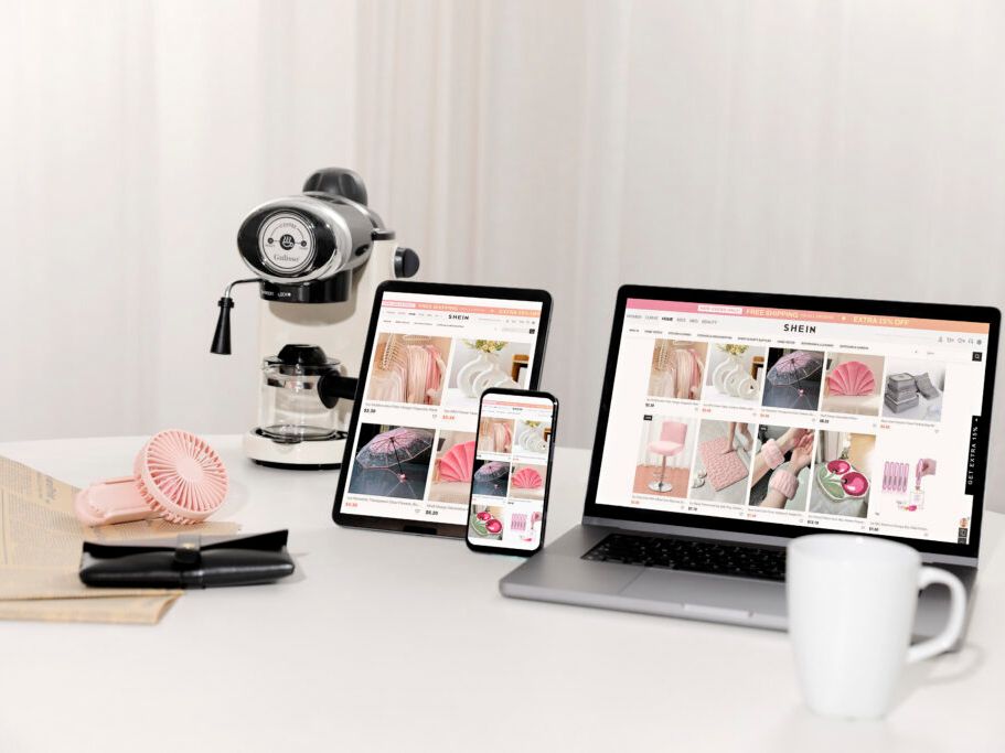 A desk setup with a coffee machine, a fan, a mug, and multiple devices (laptop, tablet, and phone) displaying the SHEIN website