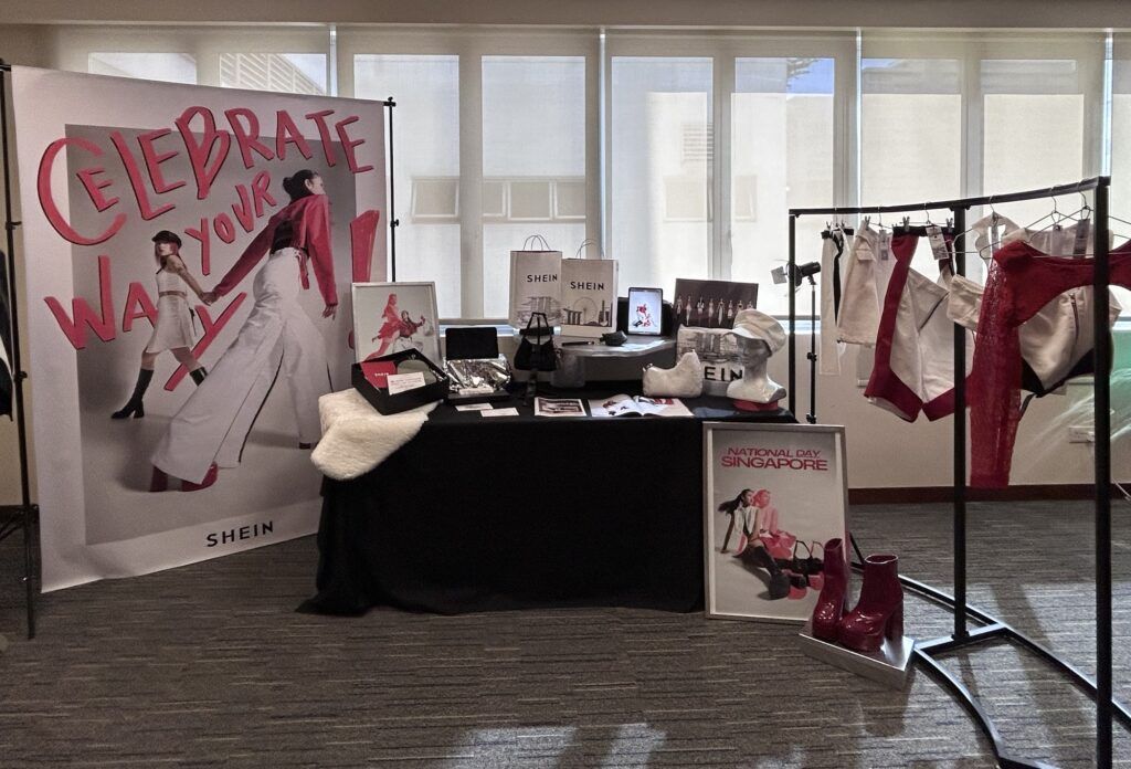 A SHEIN display features a banner, table with accessories, and clothing on a rack indoors