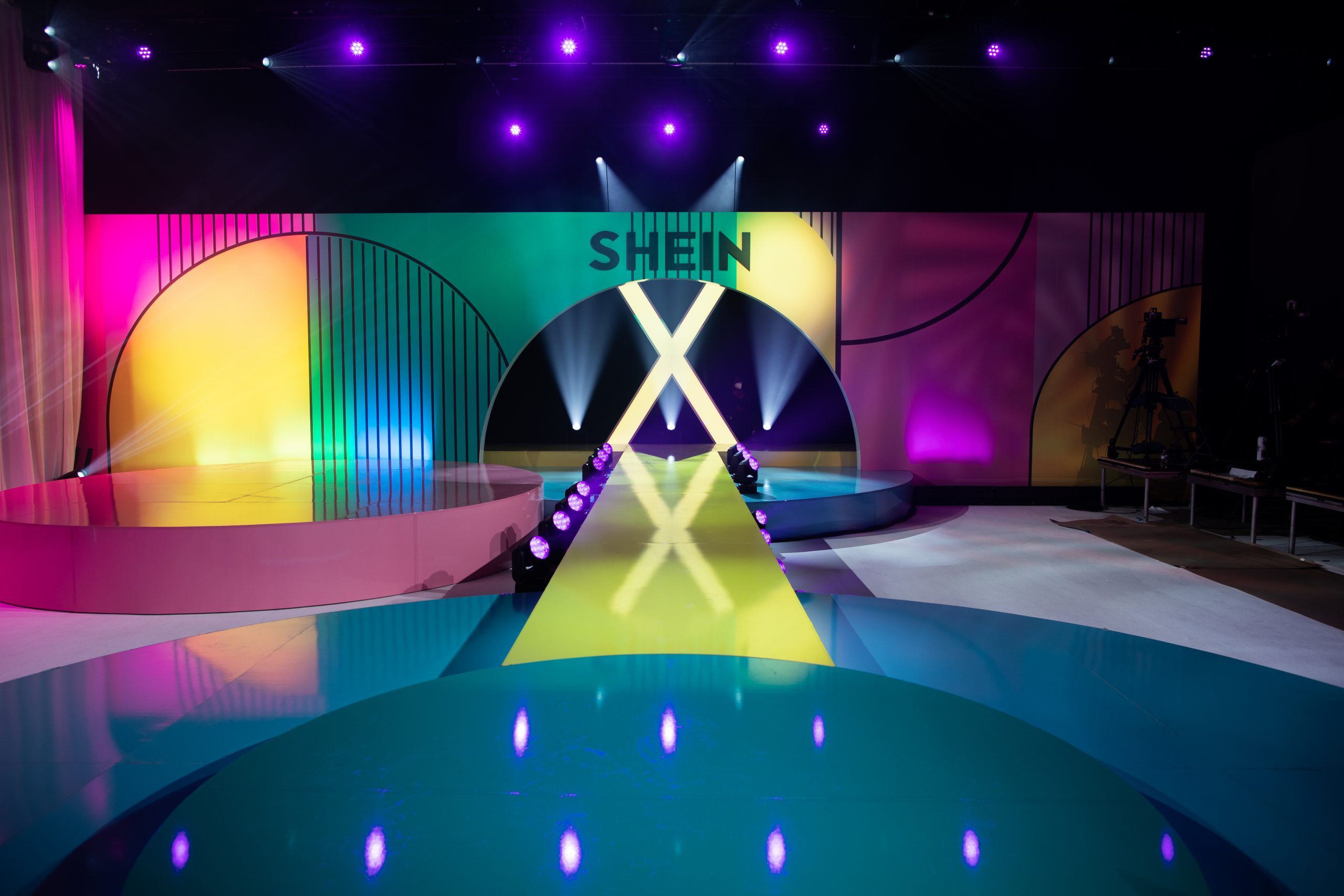 A brightly lit fashion runway stage with colorful geometric designs and "SHEIN X" displayed above the entrance