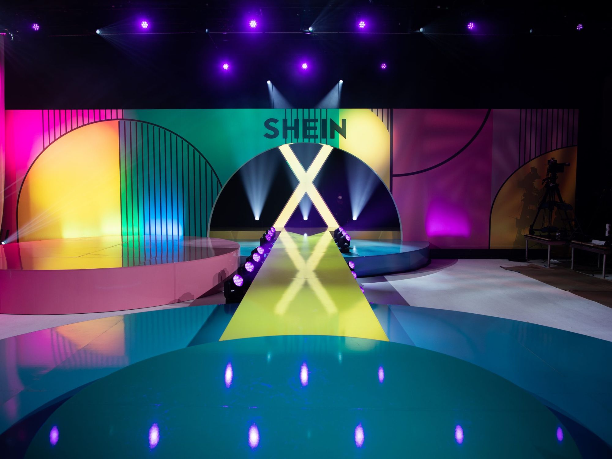 A brightly lit fashion runway stage with colorful geometric designs and "SHEIN X" displayed above the entrance