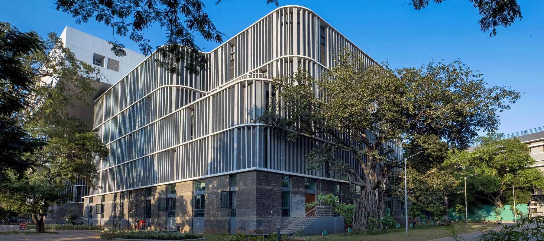 Collector Office, Pune: curved facade of vertical louvres above a stone plinth, seen through mature trees