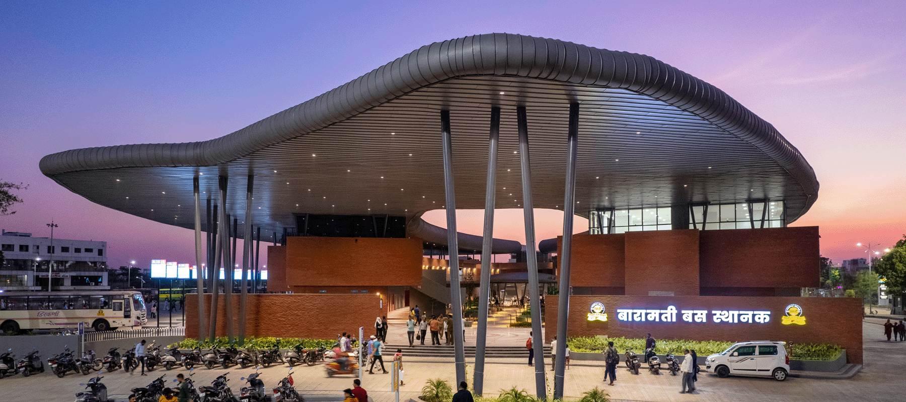 Bus Station, Baramati at dusk: an undulating steel canopy floating over brick volumes