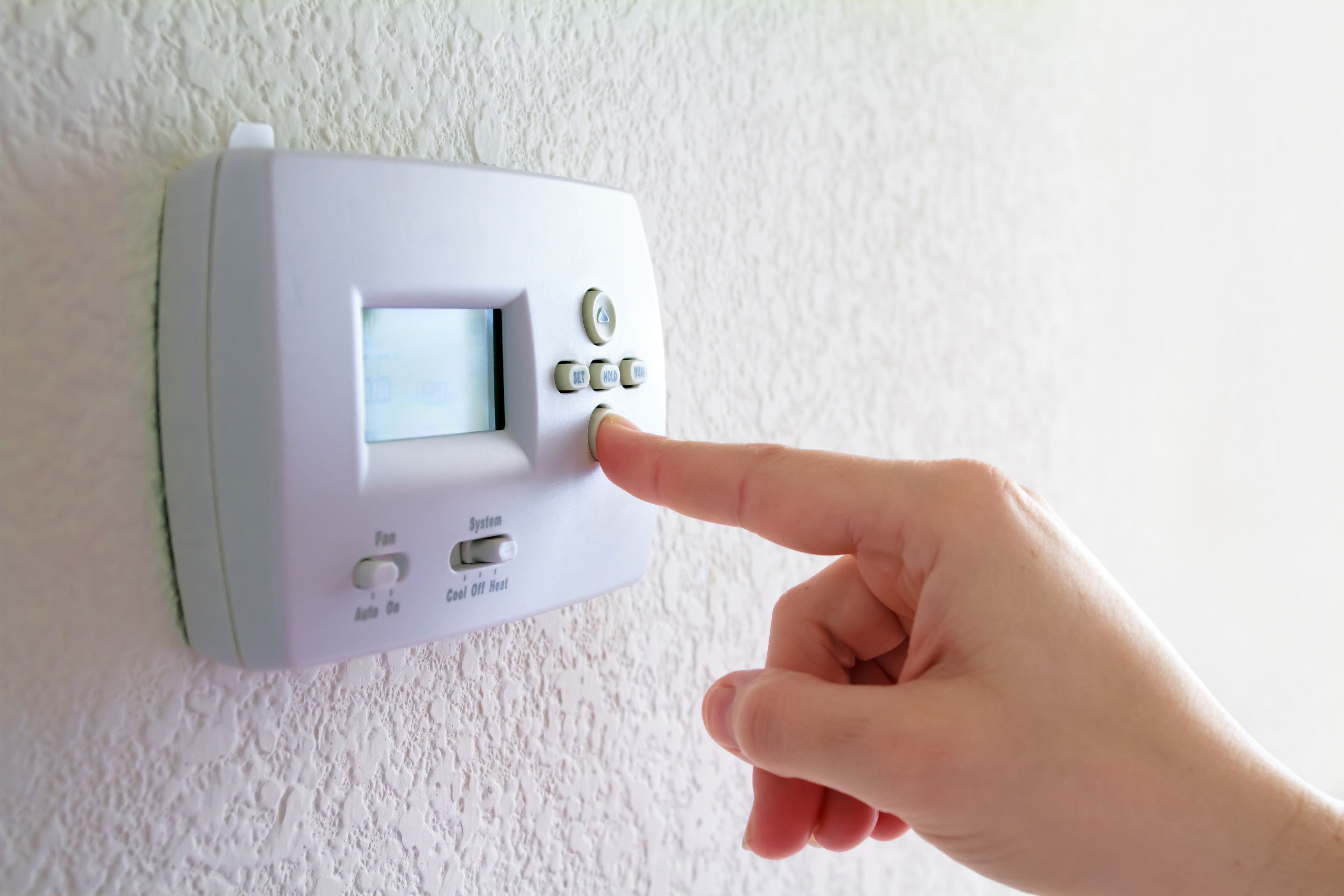 A finger pressing a button on a wall thermostat, with the Fan and System switches visible below the display.