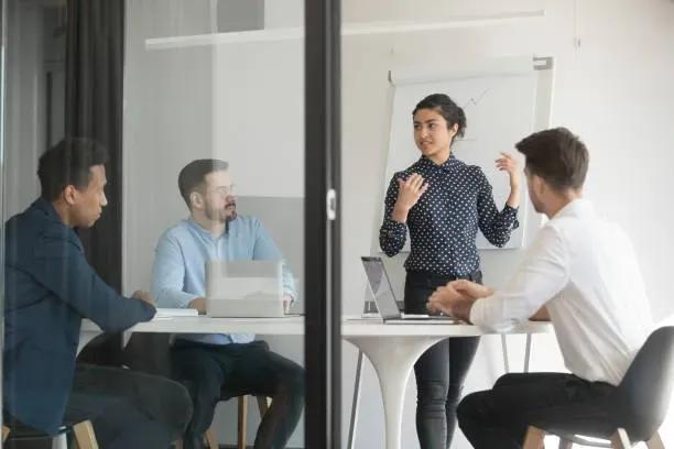 Business team in a meeting room with a woman presenting strategy on a whiteboard while colleagues listen and discuss.