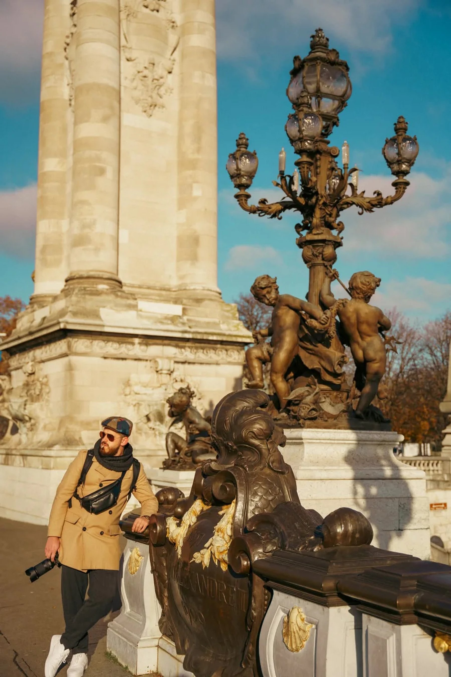 Ergys Shehu in a coat and flat cap beside an ornate stone monument and lamp post, photo 2