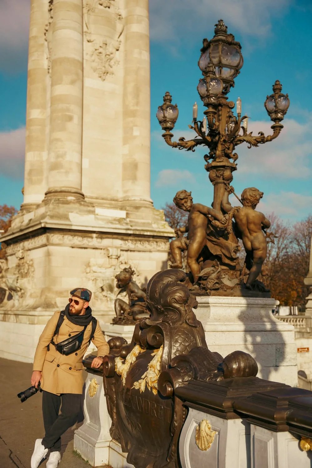 Ergys Shehu in a coat and flat cap beside an ornate stone monument and lamp post, photo 2
