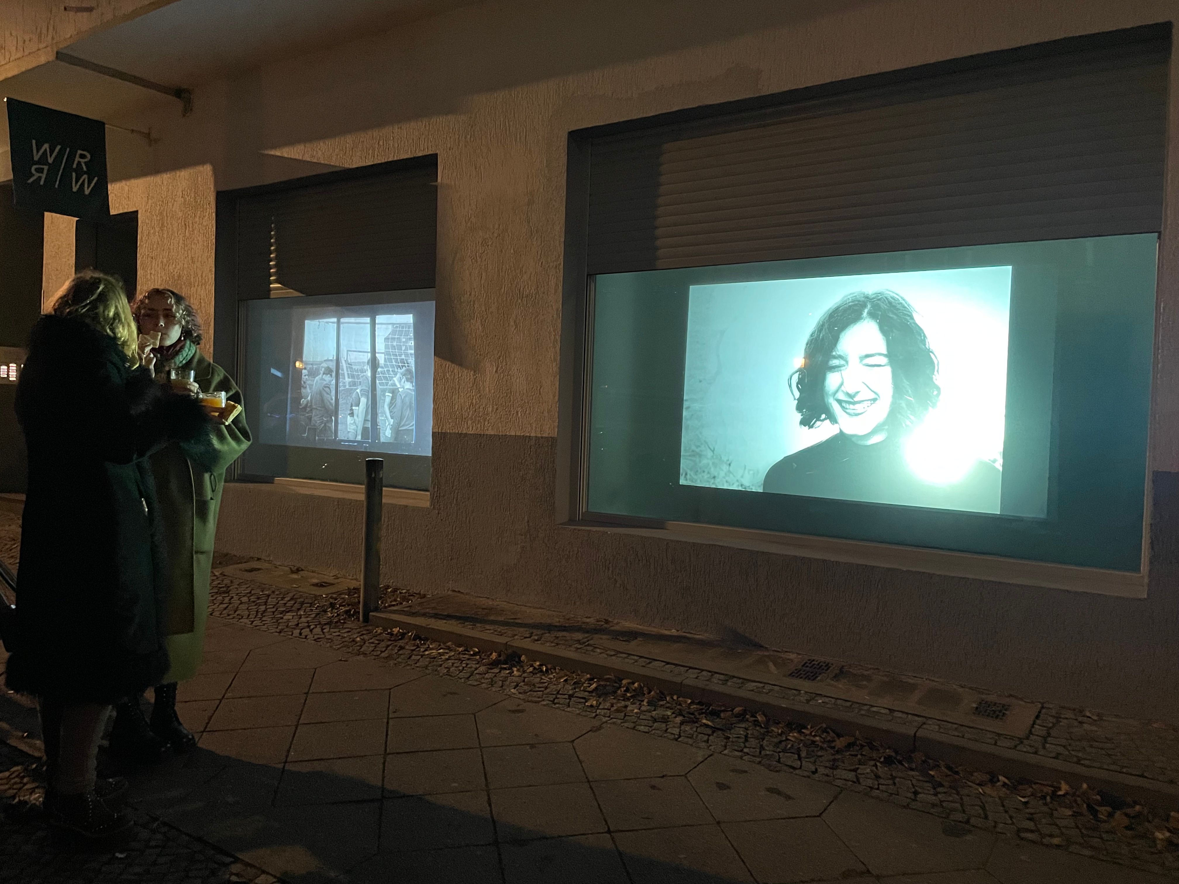 Two people stand, eating, on the curb in front of two projected photographs: a smiling portrait and people behind a window.