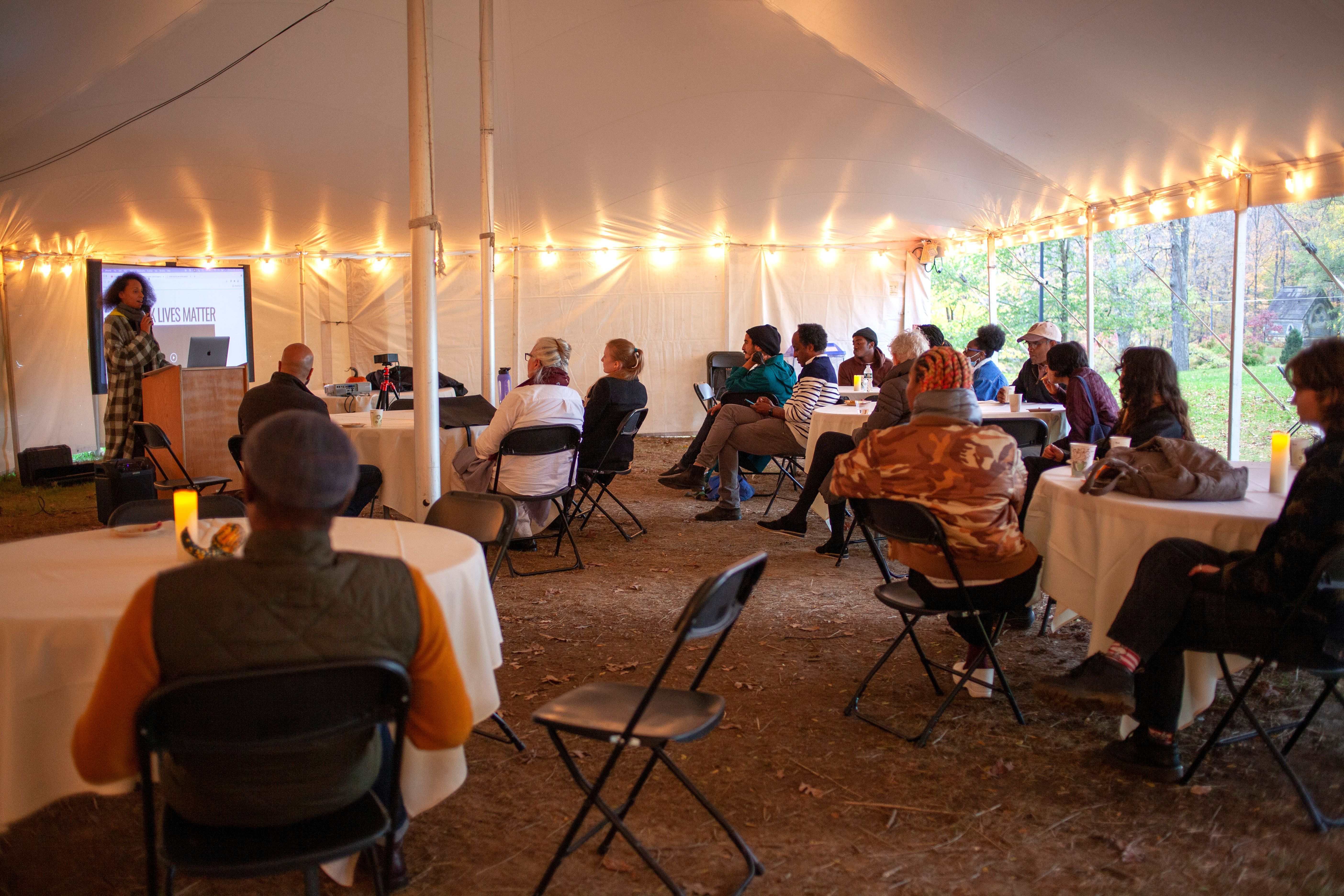 People under a white tent at white-clothed, circular tables sit, watching a presenter speak (Powerpoint slide reads: Black Lives Matter)