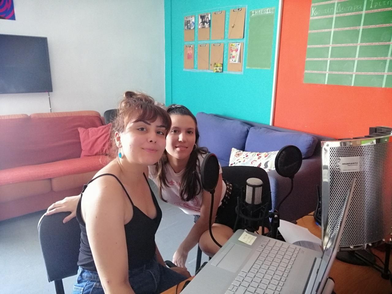 Two students smile in front of a laptop and podcasting microphone