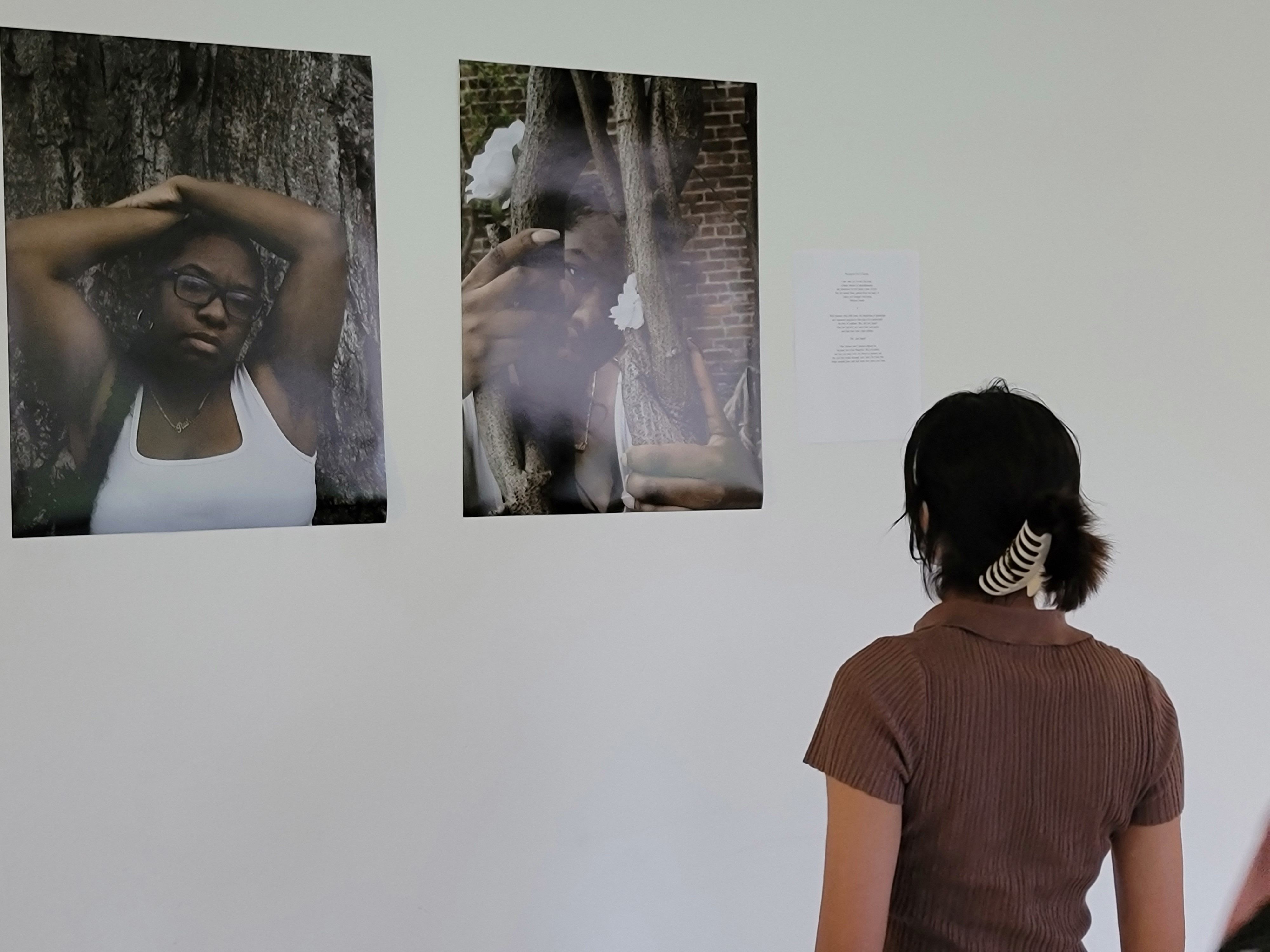 A person looks at a wall at two poster-sized prints: self-portraits of artist Rasheeda Graham in front of a tree trunk and peering through twisting branches.