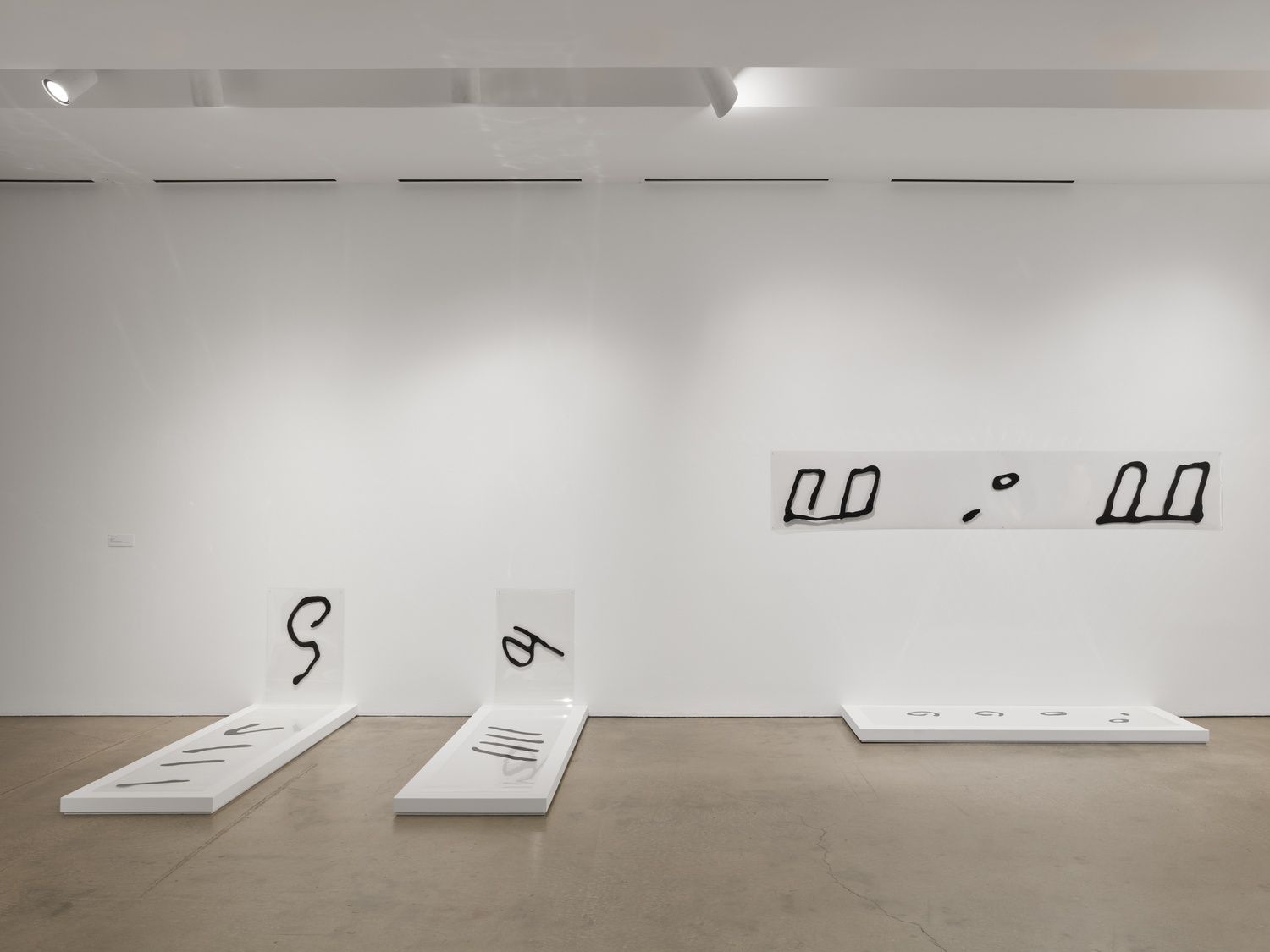 A white wall and three white, rectangular slabs on the floor. The slabs and three sections of the wall are painted with simple, black symbols (all abstract: circles and lines, a hook shape).
