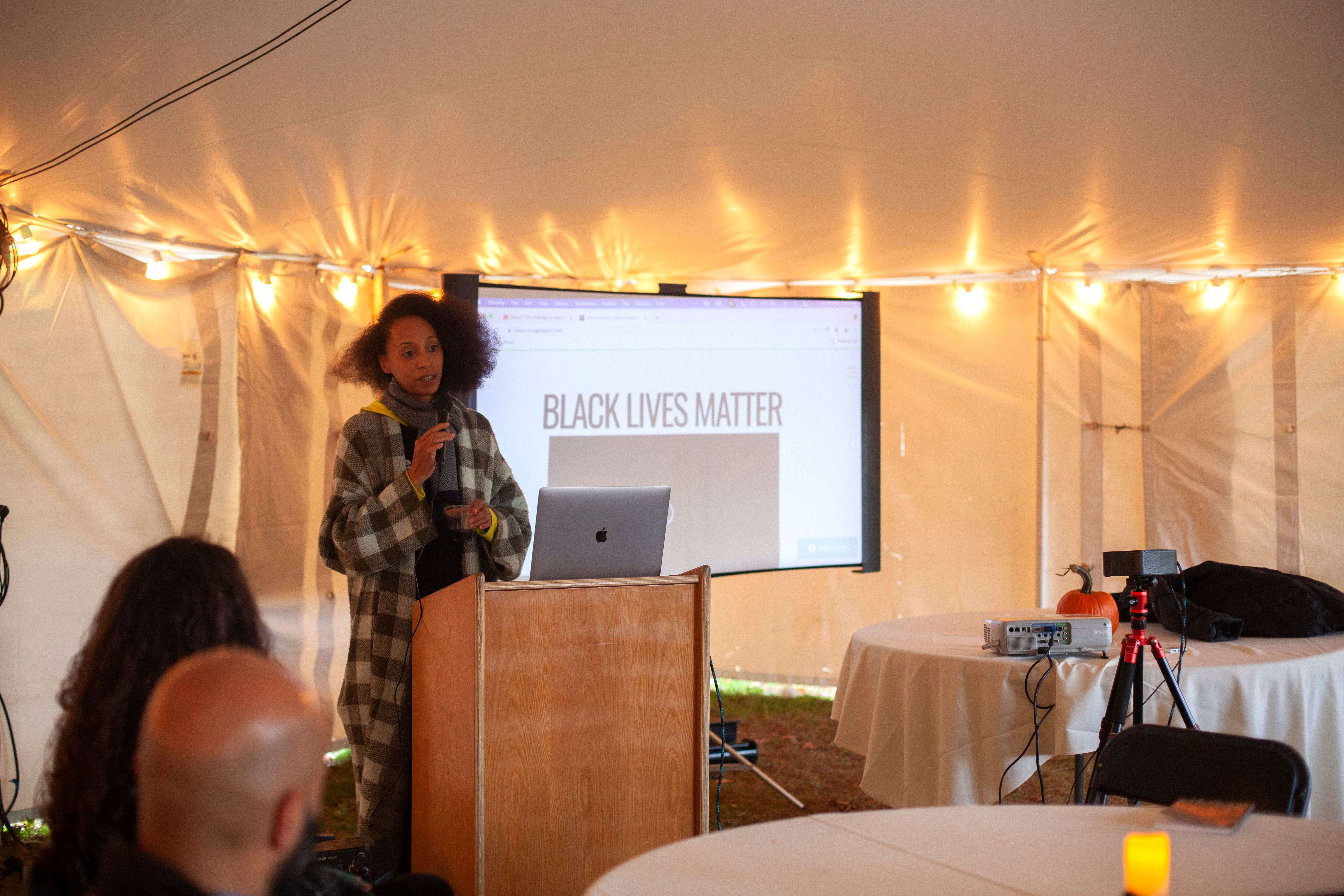 A presenter speaks at a podium in front of a powerpoint slide reading “Black Lives Matter”