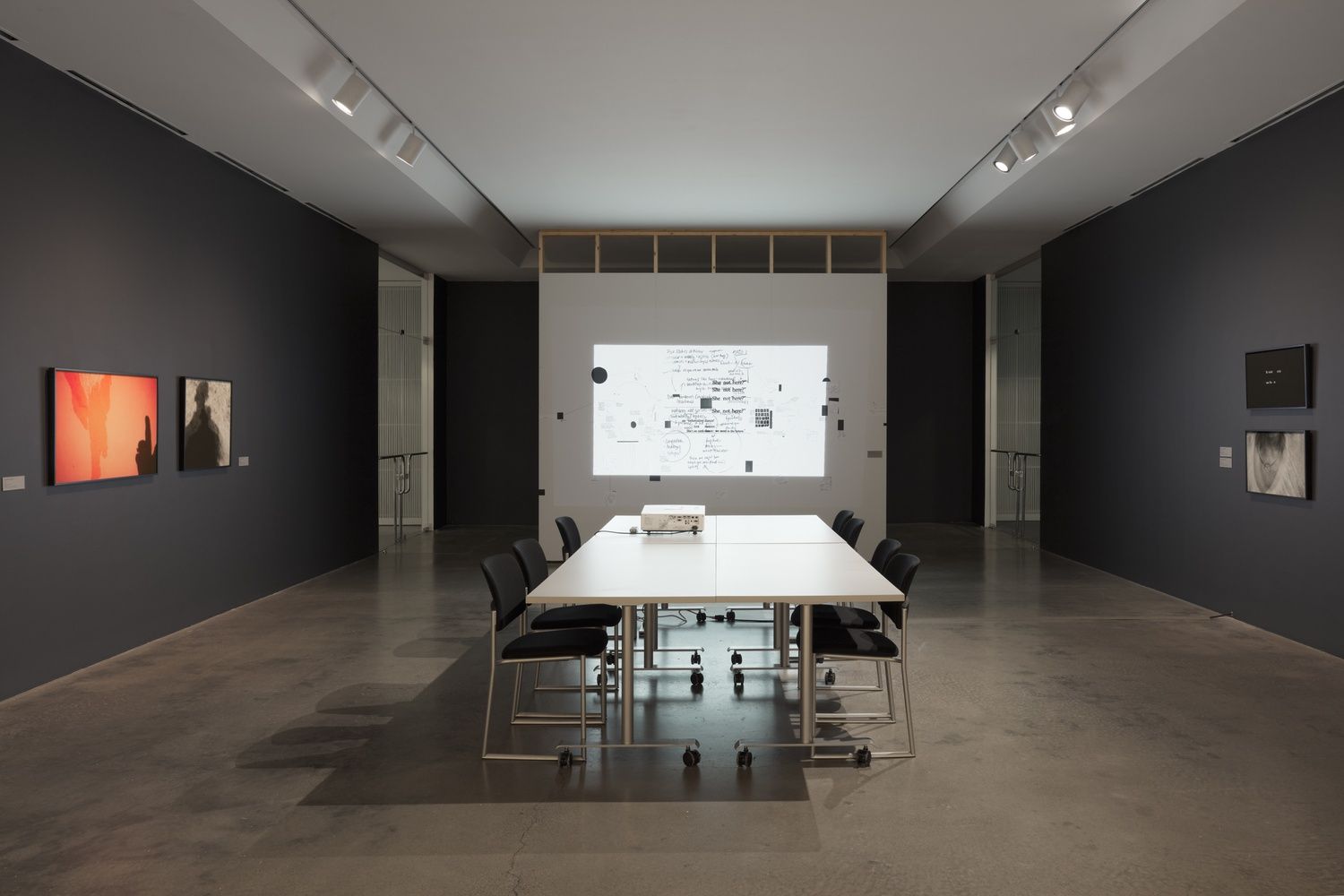 A long room with a projector on a conference table with chairs. It projects a white screen with black notes and geometric shapes. On the walls are prints of shadowy figures in black and white and orange.