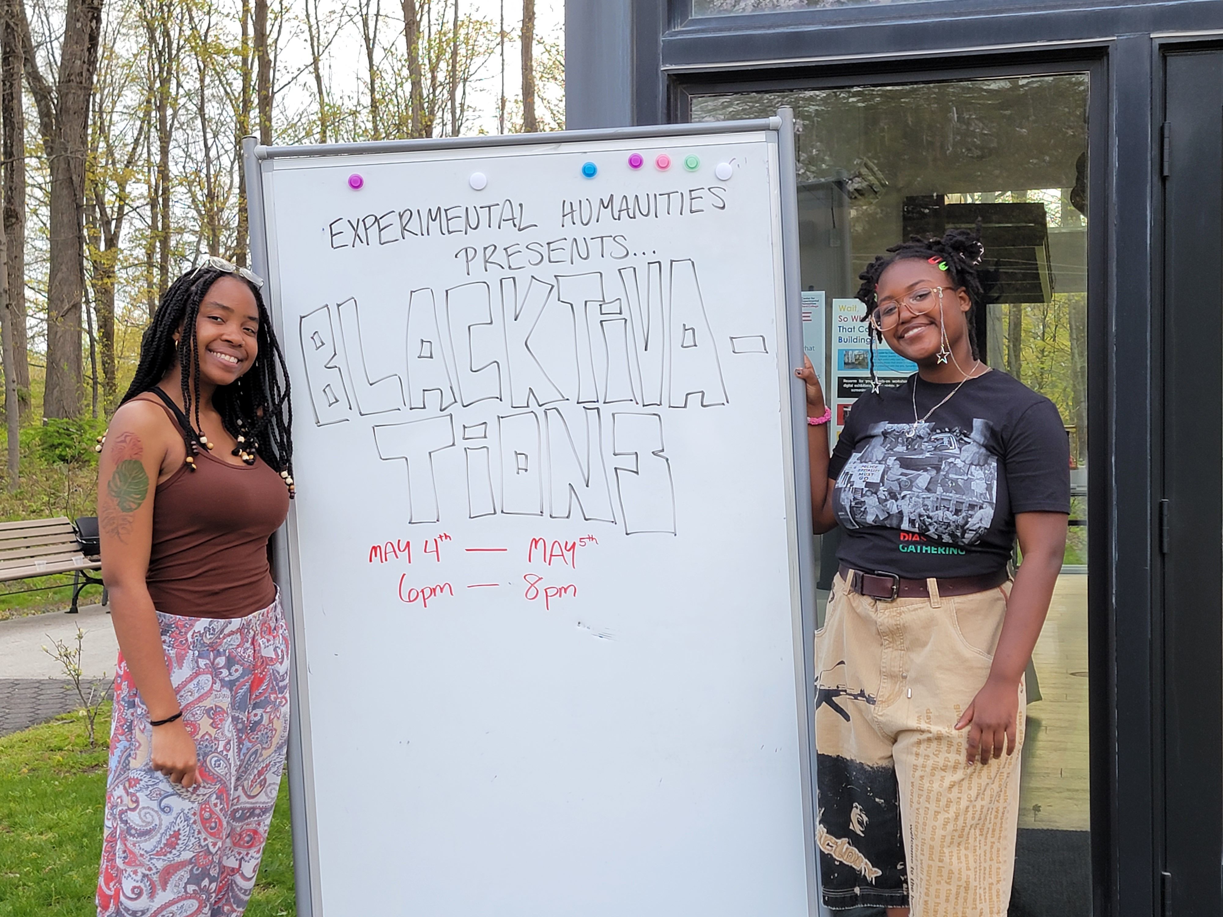 Two Black Imagination student artists with a whiteboard that reads: Experimental Humanities Presents: Blacktivations May 4th- May 5th 6pm-8pm.